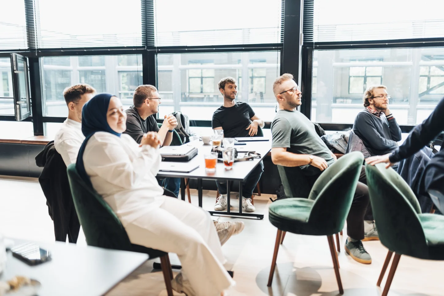 Workshop attendees seated around tables beside large windows, listening to a speaker.